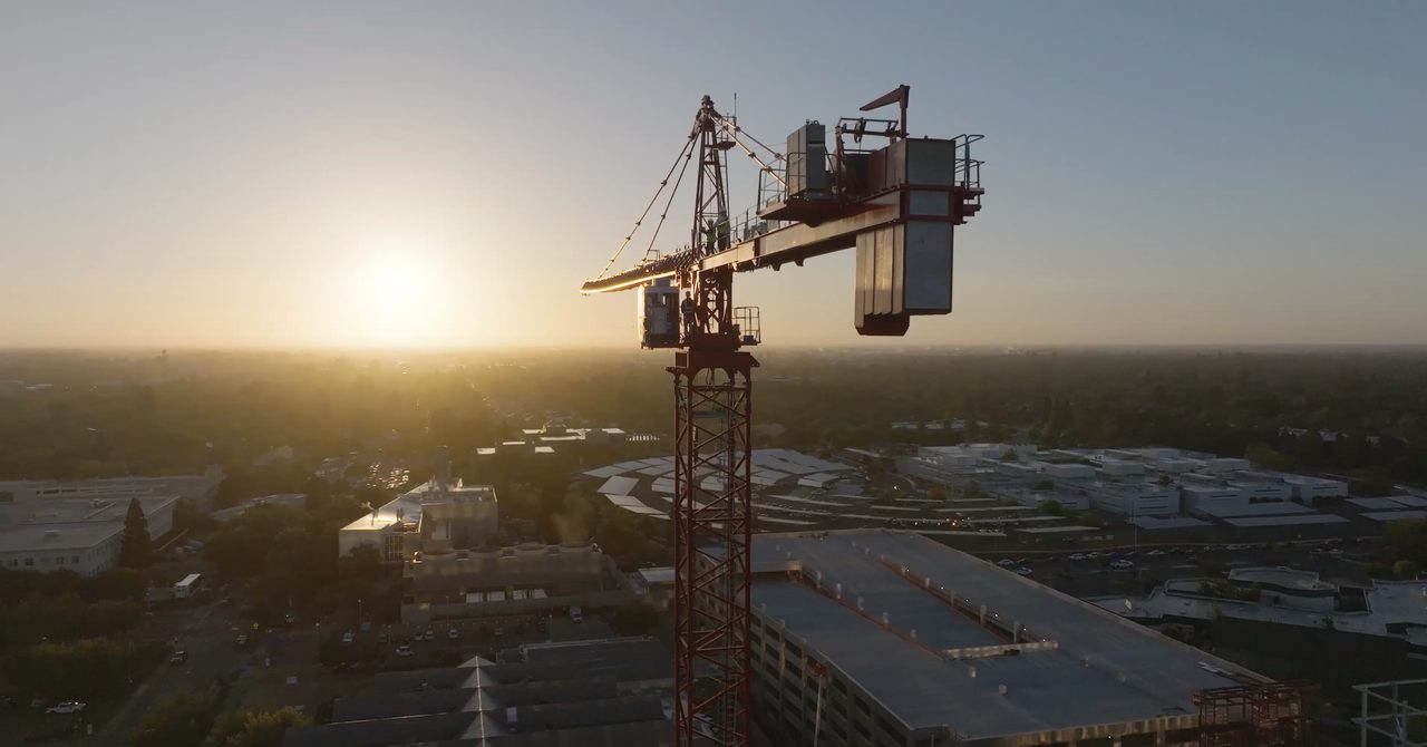 A tower crane silhouetted against the sunrise over the Aggie Square construction site, crew members on the jib.