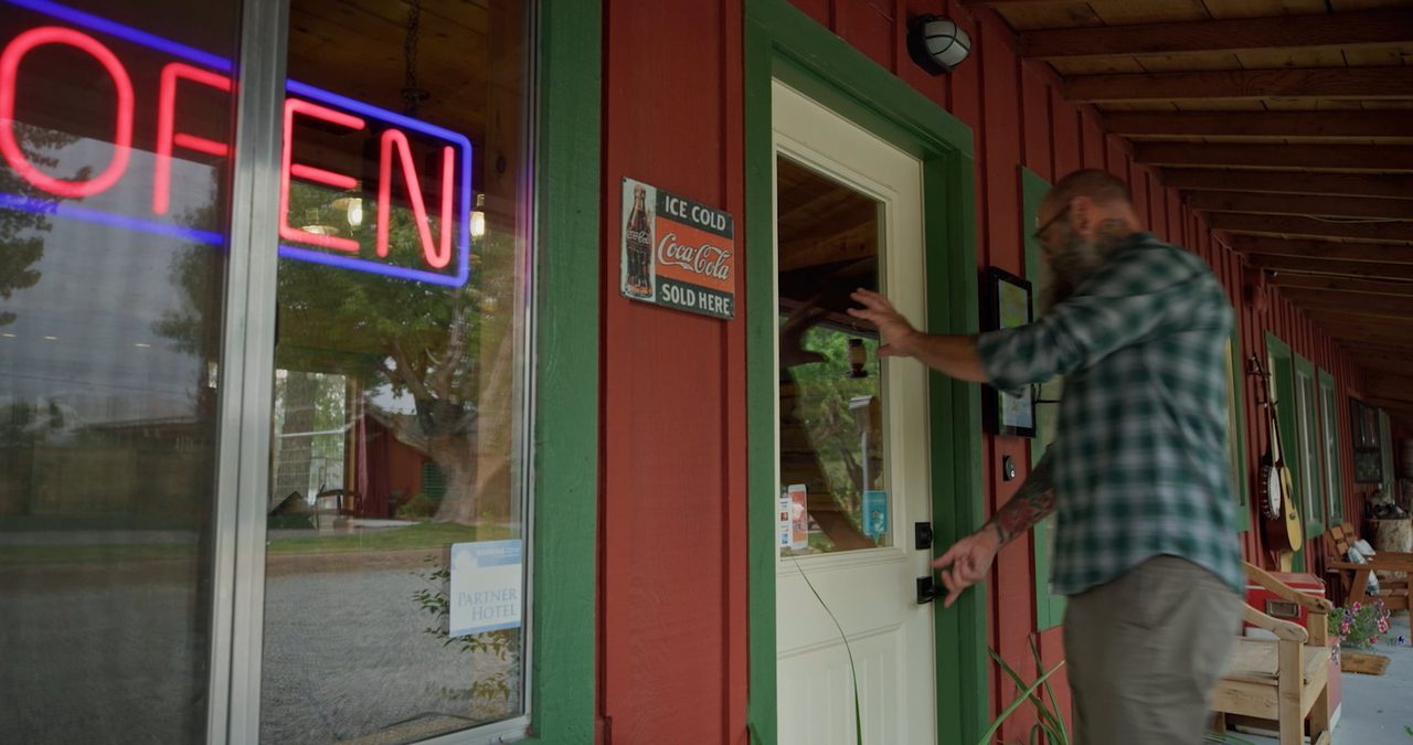 Steve opens the front door of the Andruss Motel beneath a glowing neon Open sign.