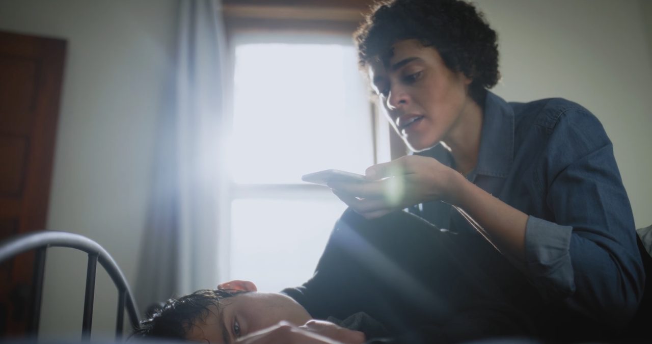 A woman leans over a young man lying on a bed, speaking urgently into her phone.