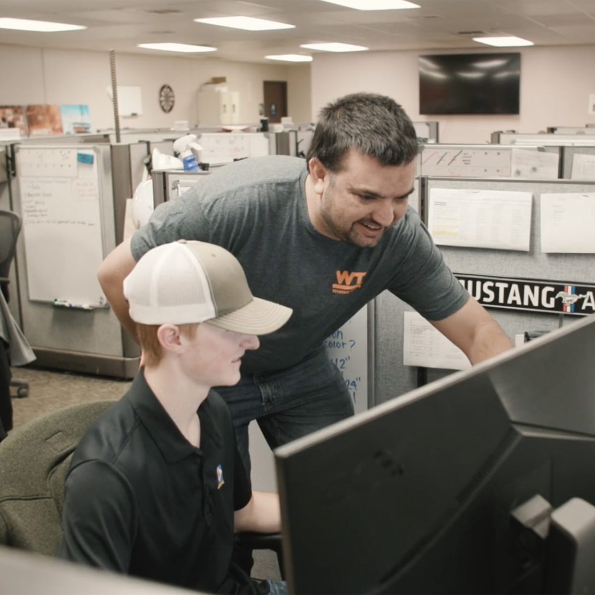 A new hire at his desk, smiling, as a teammate leans in to look at his screen with him.